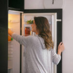 Happy woman opening the fridge and taking food from the upper shelf